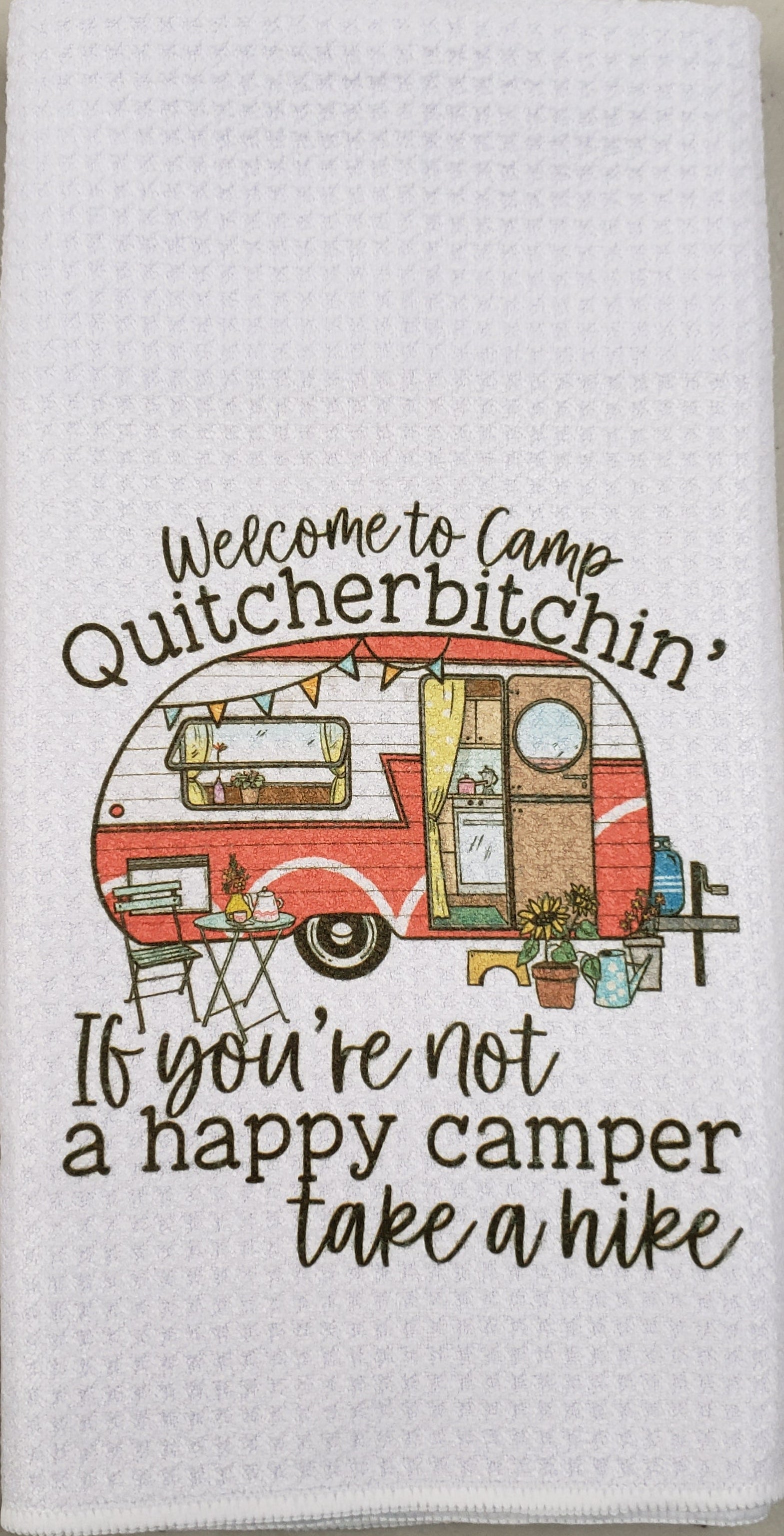 A white waffle-weave kitchen towel featuring a retro red and orange camper illustration with the text 'Welcome to Camp Quitcherbitchin'' and 'If you're not a happy camper take a hike'.